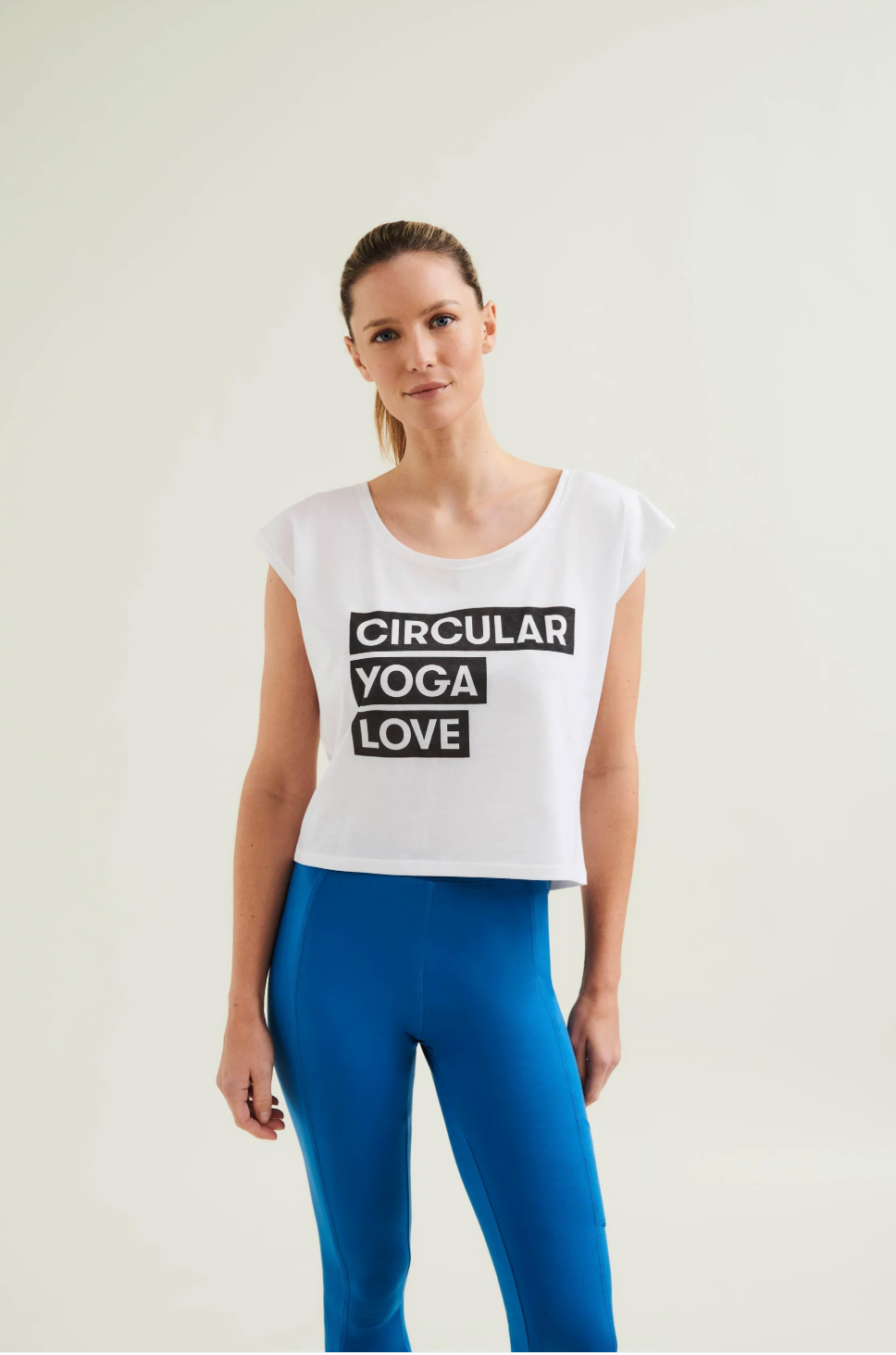 Woman wearing a white tank top with 'Circular Yoga Love' text and blue leggings on a light background