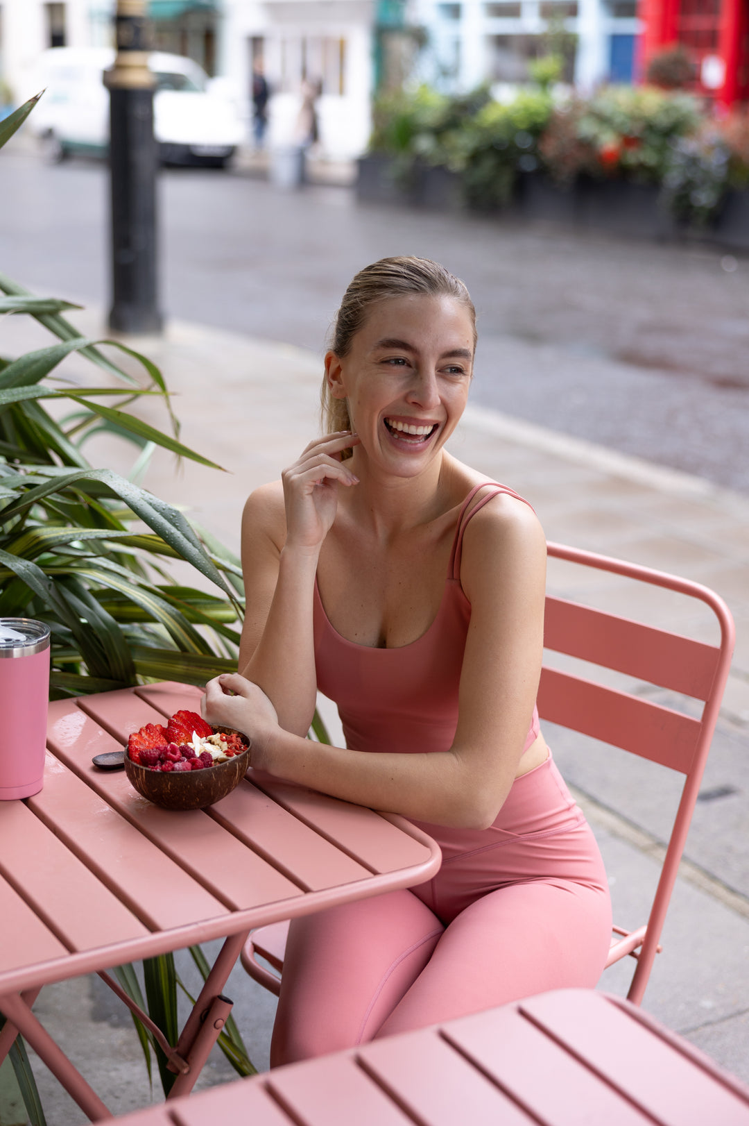 Woman in pink activewear outfit sitting at a pink table outdoors, laughing.