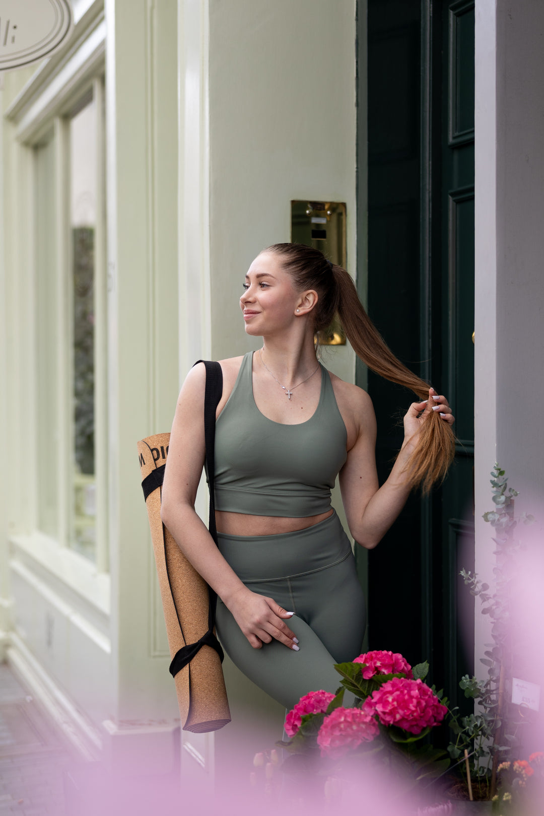 Woman in green activwear standing in a doorway carrying Warrior Addict's cork yoga mat  with flowers and a chair in the foreground.