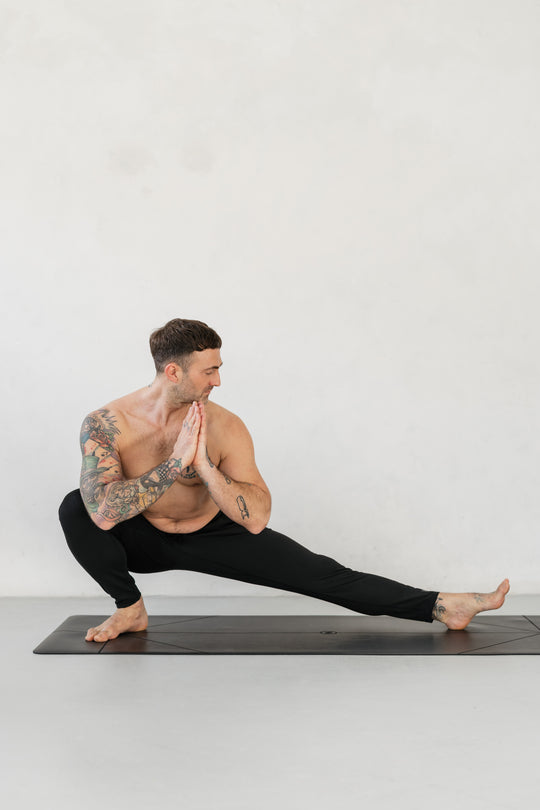 Man in yoga pose on mat. Tattoos on arms and legs. Black pants. Hands in prayer position. White background.