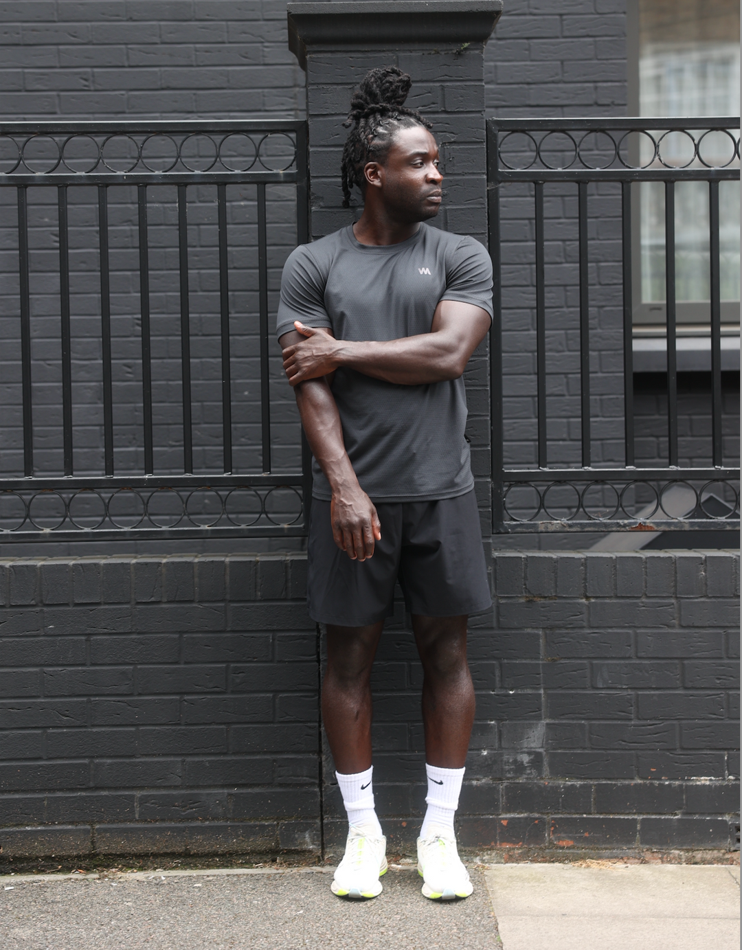 Man in black activewear by Warrior Addict standing against a black brick wall with a metal fence.