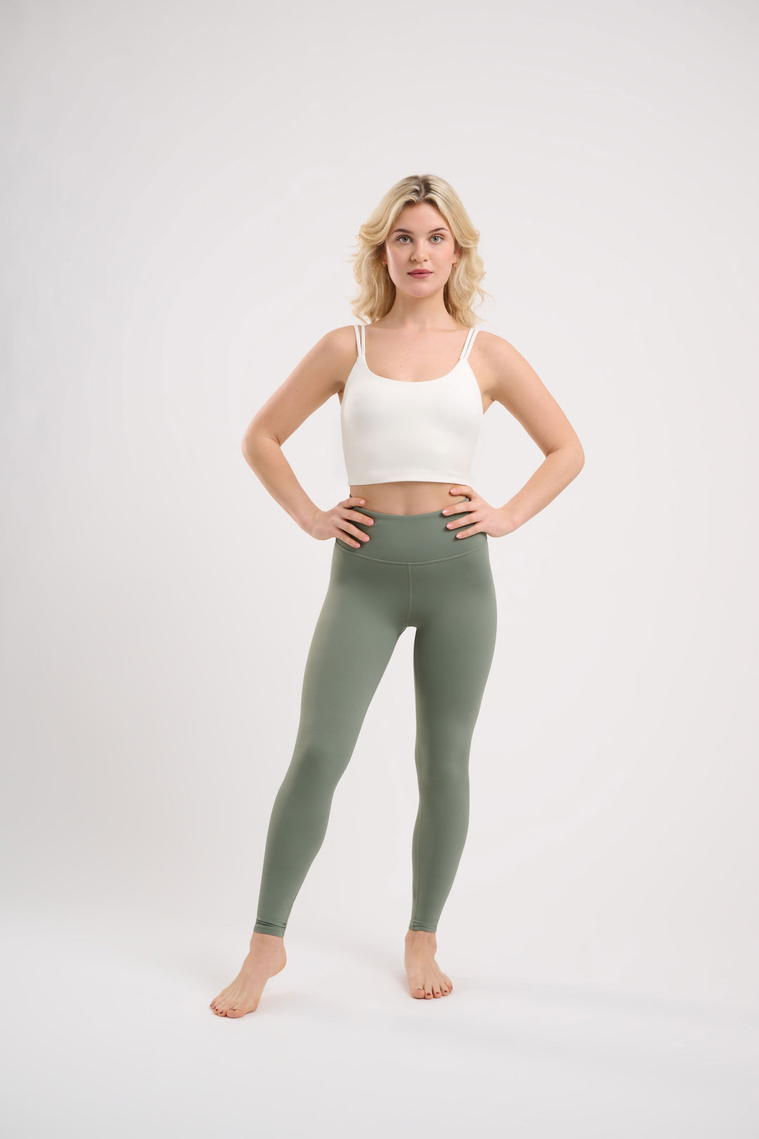 A blonde woman stands with hands on hips, wearing a white crop top and green leggings.