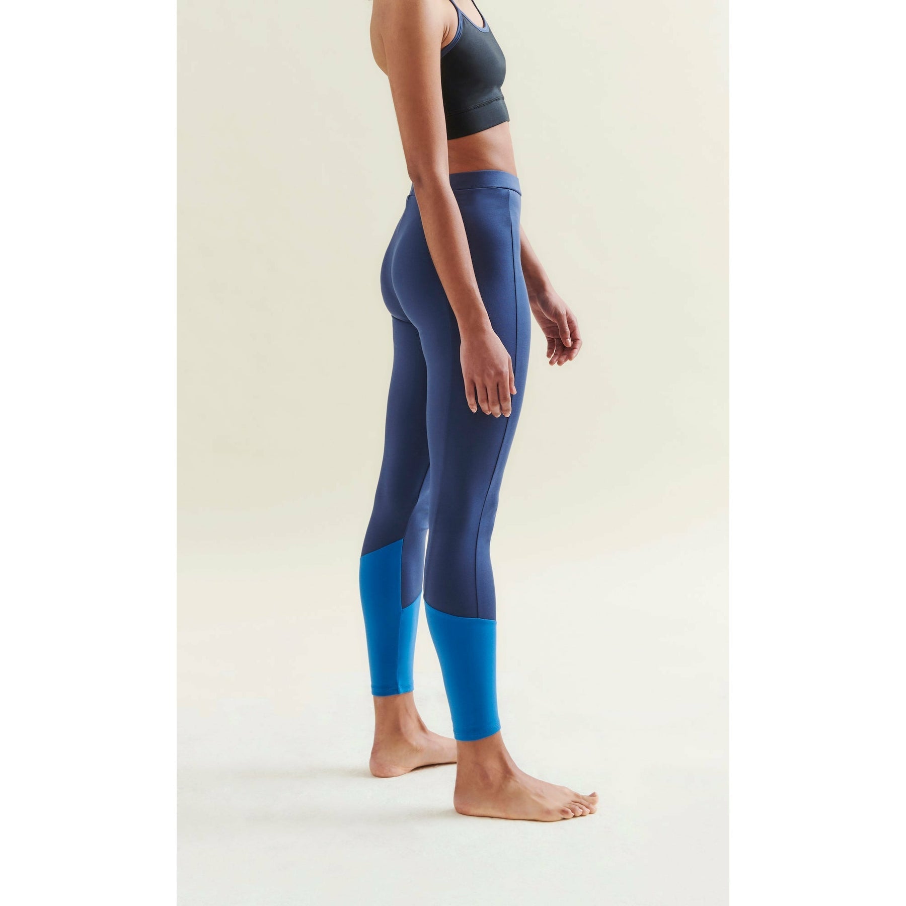 Woman wearing a black sports bra and two-tone blue leggings, barefoot in a studio setting.
