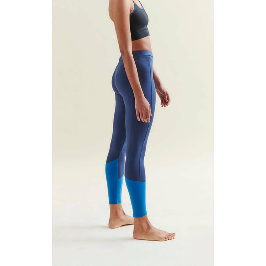 Woman wearing a black sports bra and two-tone blue leggings, barefoot in a studio setting.