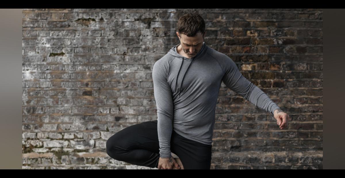 man wearing lomg sleeved yoga top in grey against a brick wall