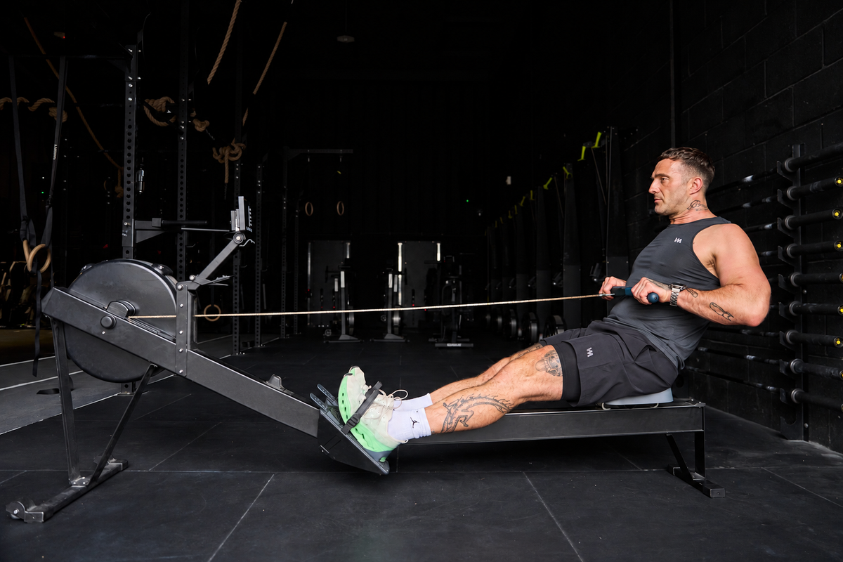 A man exercising on a rowing machine in a dark gym, wearing a black warrior addict tank top, black  warrior addict AG shorts, white socks, and neon green sneakers.