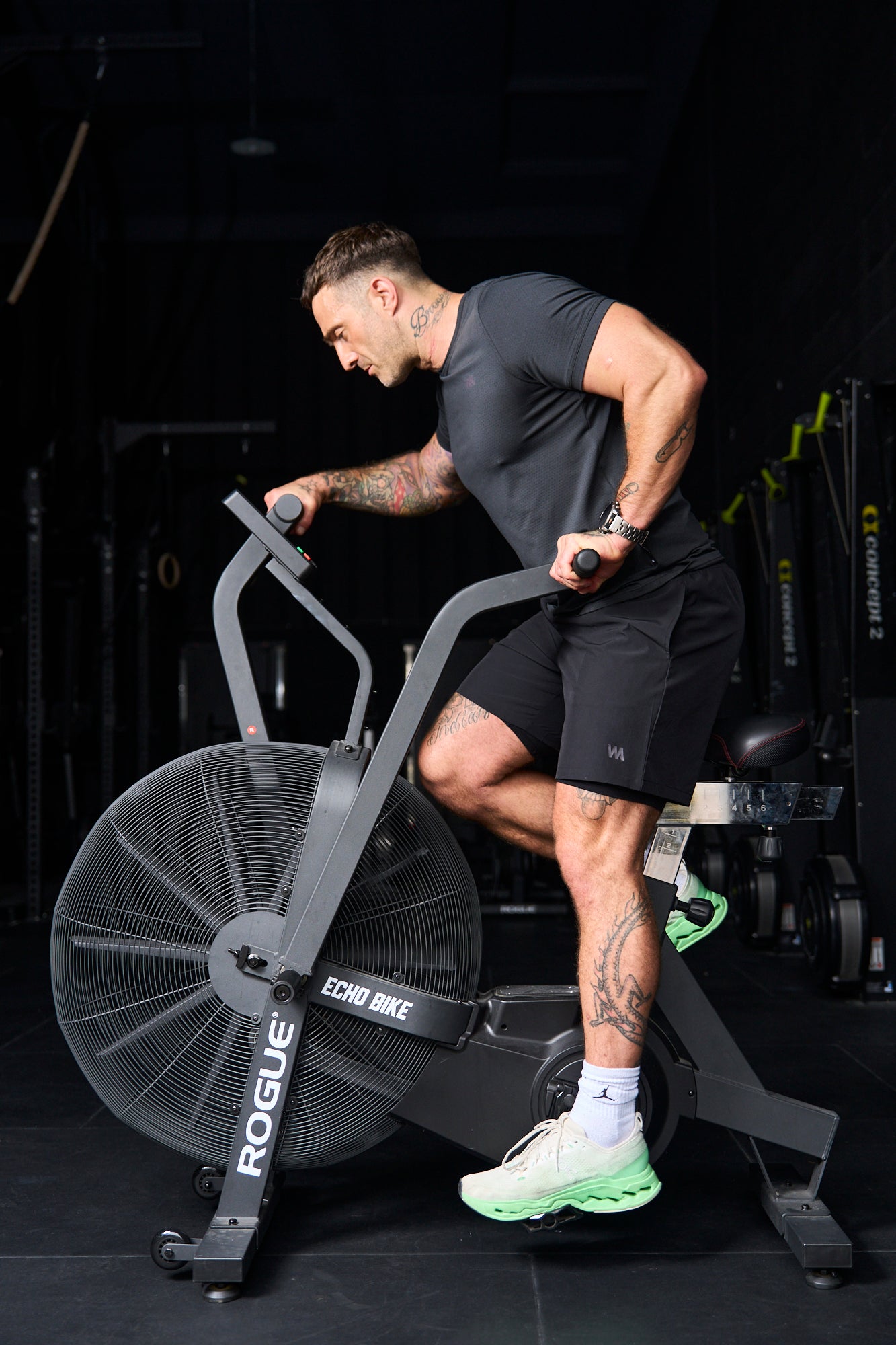 Man exercising on a Rogue stationary bike in a gym setting wearing a bkack warrior addict t-shirt and shorts 