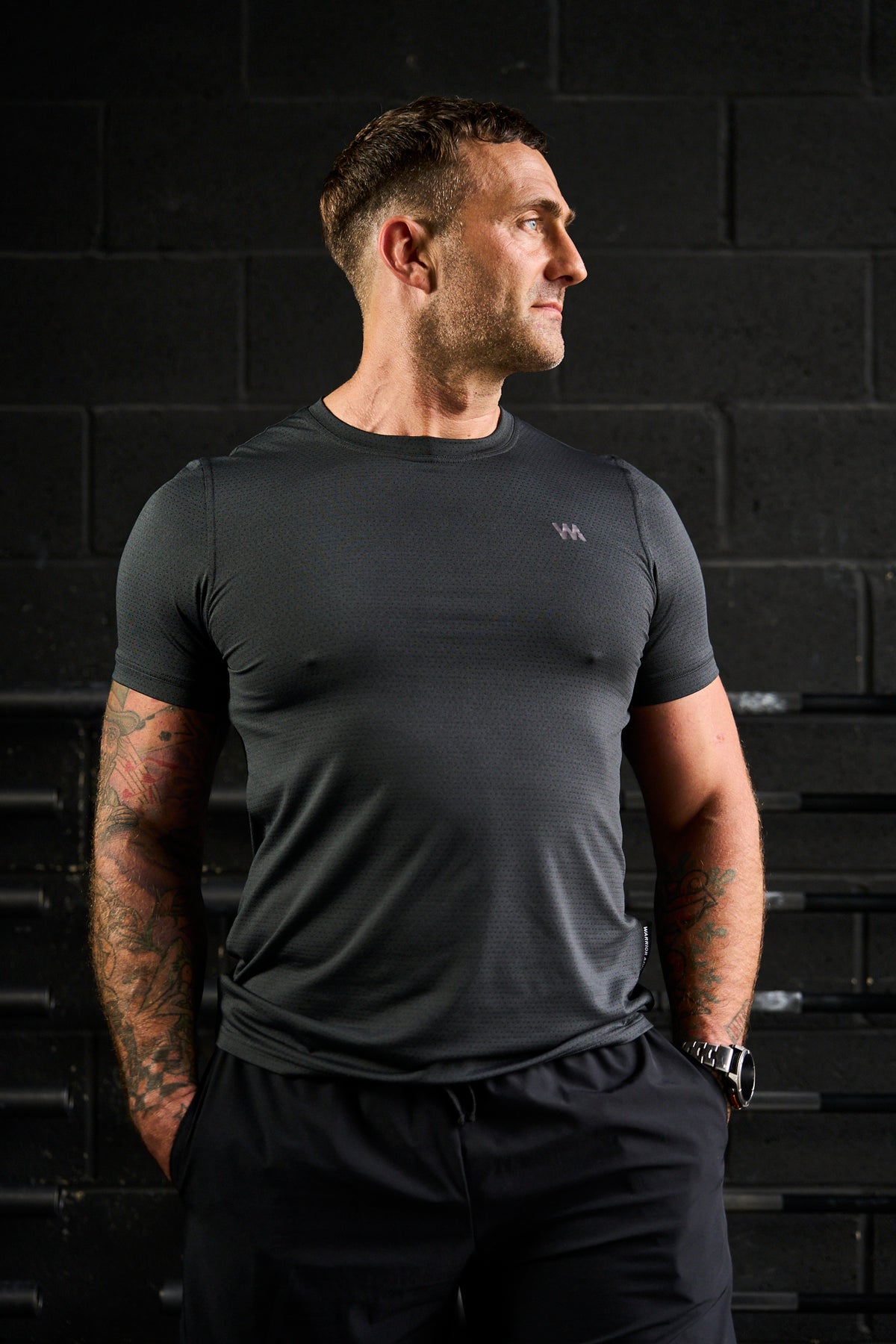 Man wearing a black  warrior addict performance t-shirt and shorts against a dark brick wall