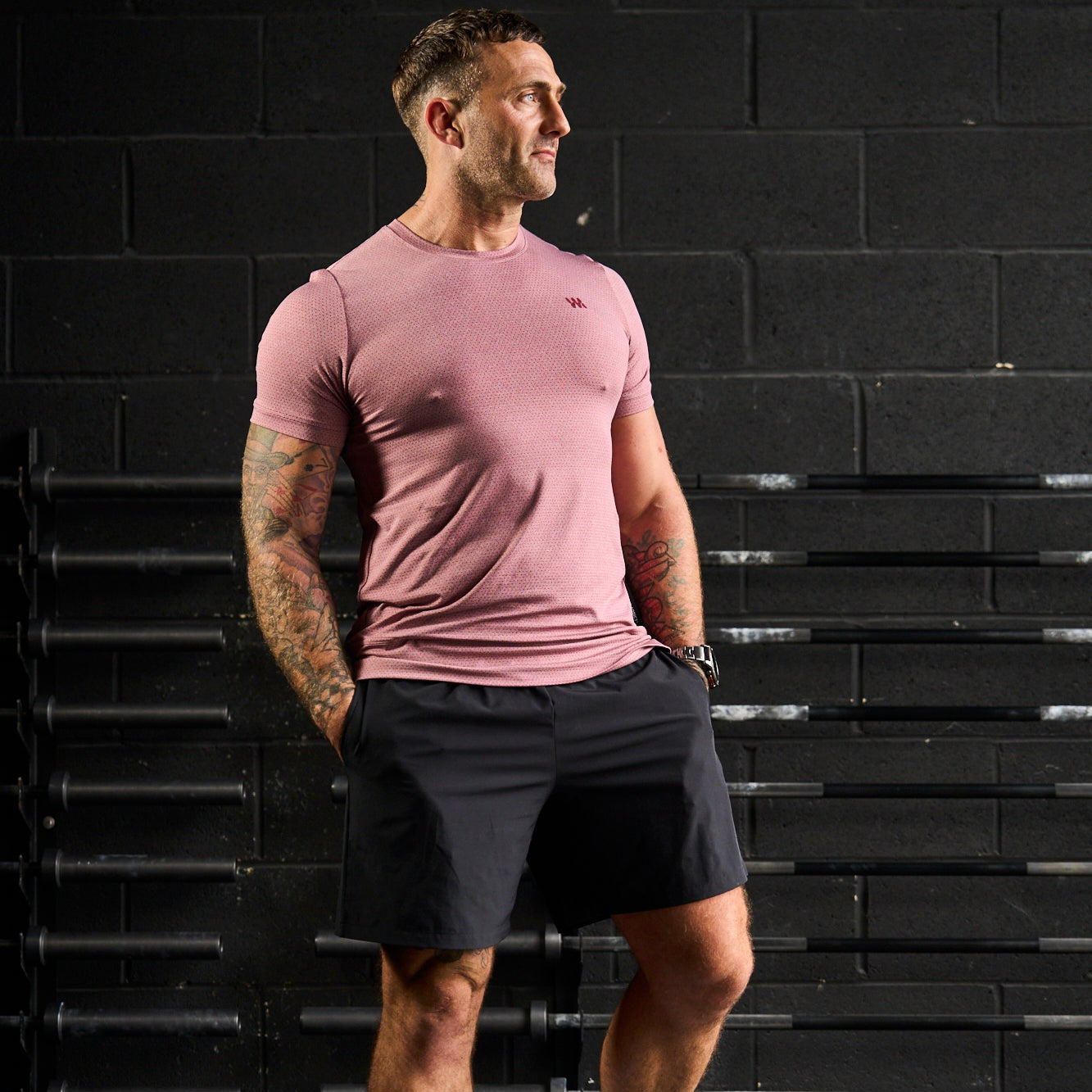 Man wearing a pink t-shirt and black shorts standing against a black brick wall.