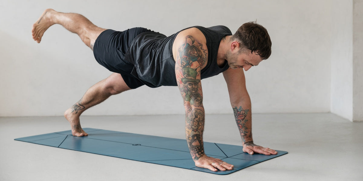 A man with tattoos performs a yoga pose on a blue yoga mat in a minimalist studio with white walls and a light gray floor.