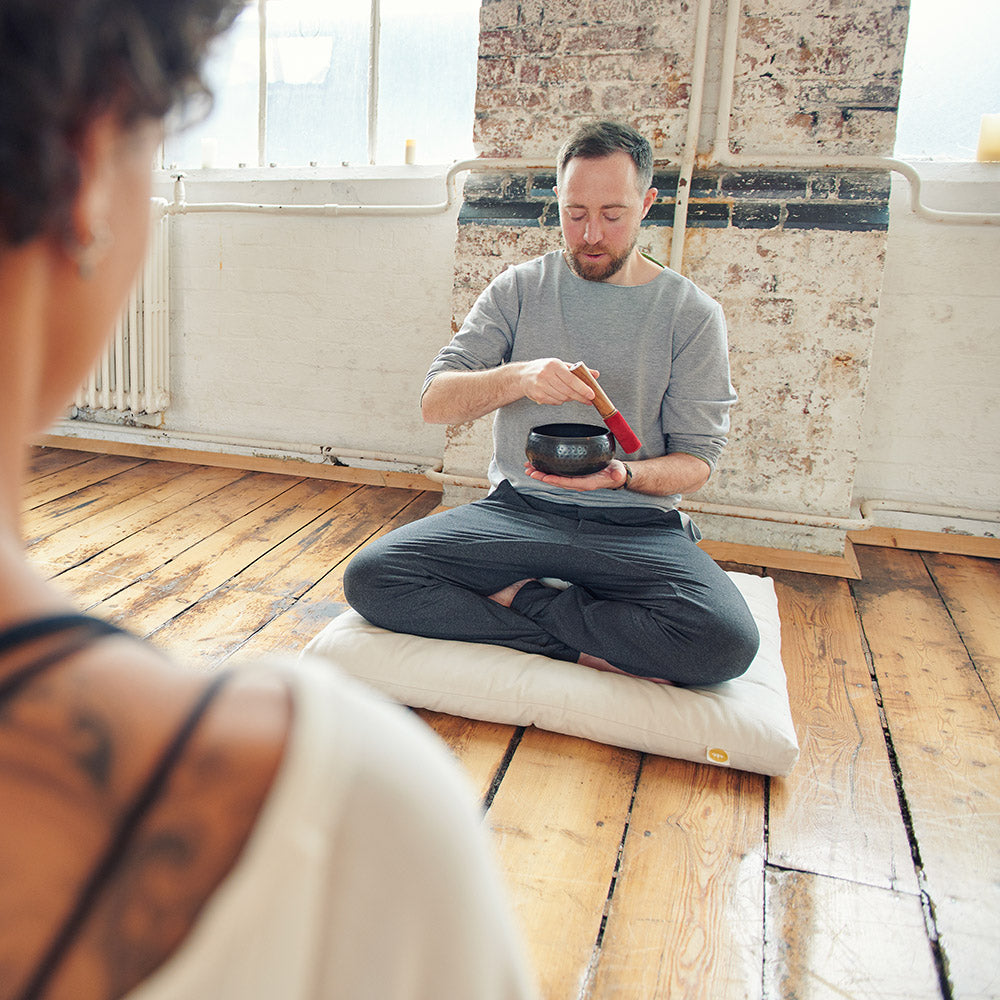 man ryan nell meditating sat crossed legged using a sound bowl in front of woman 